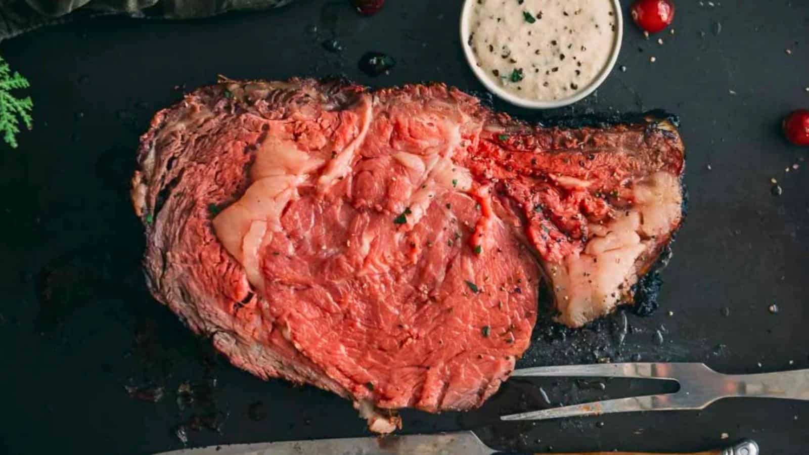 A slice of rare prime rib on a dark surface with a cup of creamy sauce and a meat fork nearby.