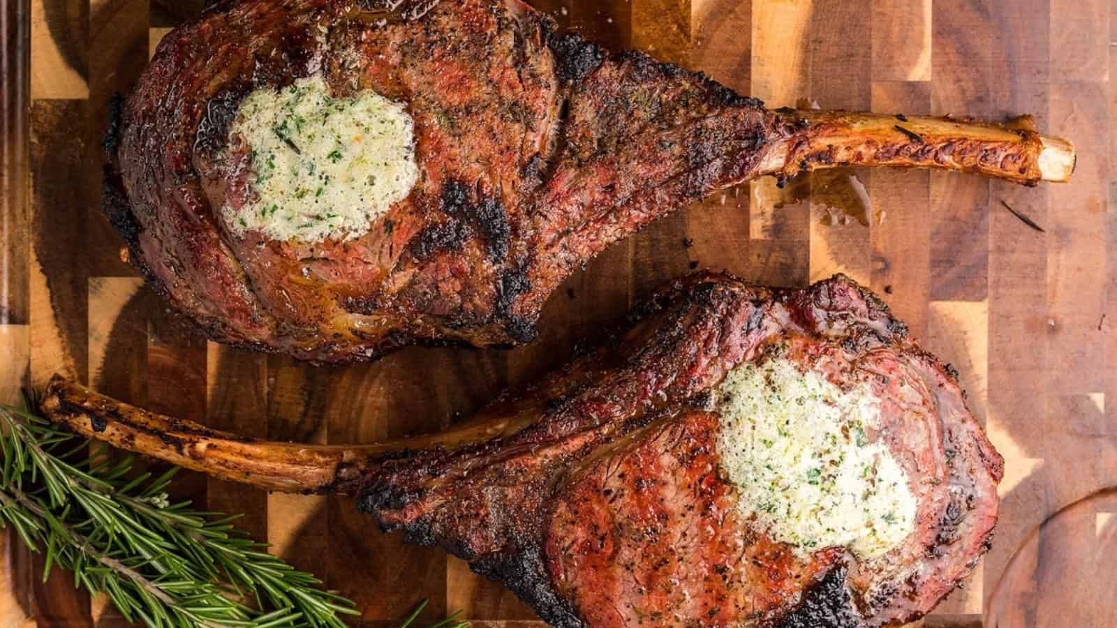 Two grilled tomahawk steaks with herb butter on top, served on a wooden board with rosemary garnish.