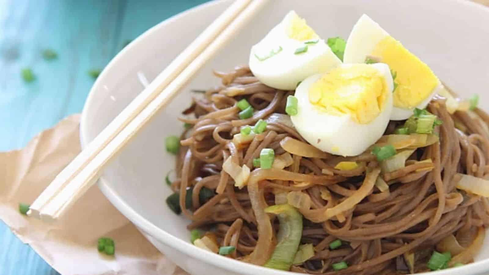 A bowl of soba noodles topped with chopped green onions and sliced boiled eggs, with chopsticks on the side.