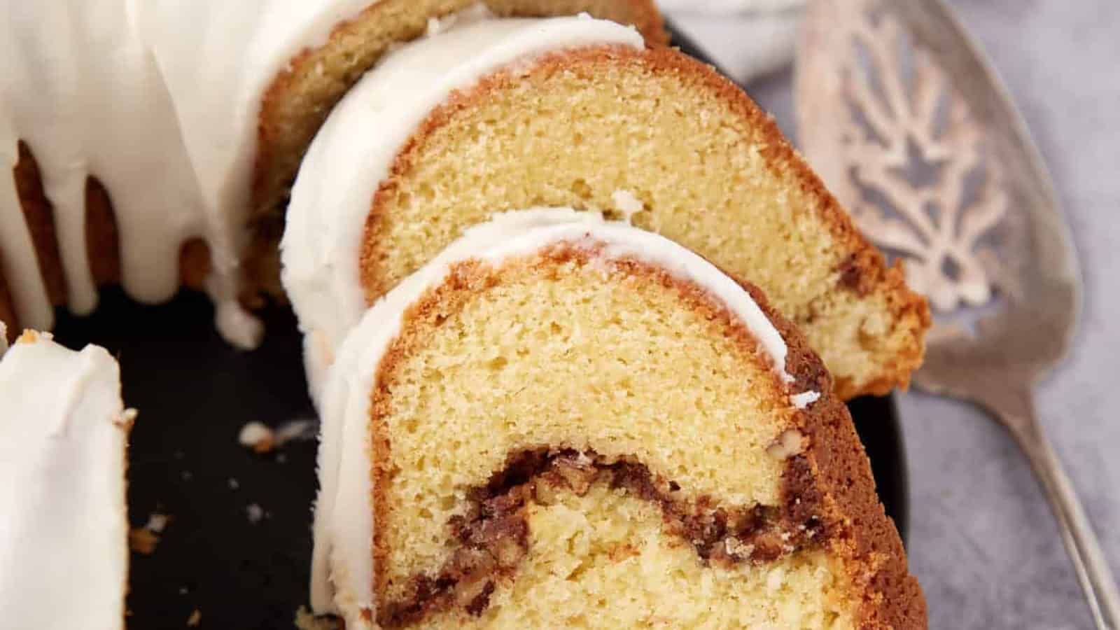 Sliced bundt cake with white icing and a cinnamon swirl, served on a black plate with a vintage cake server.