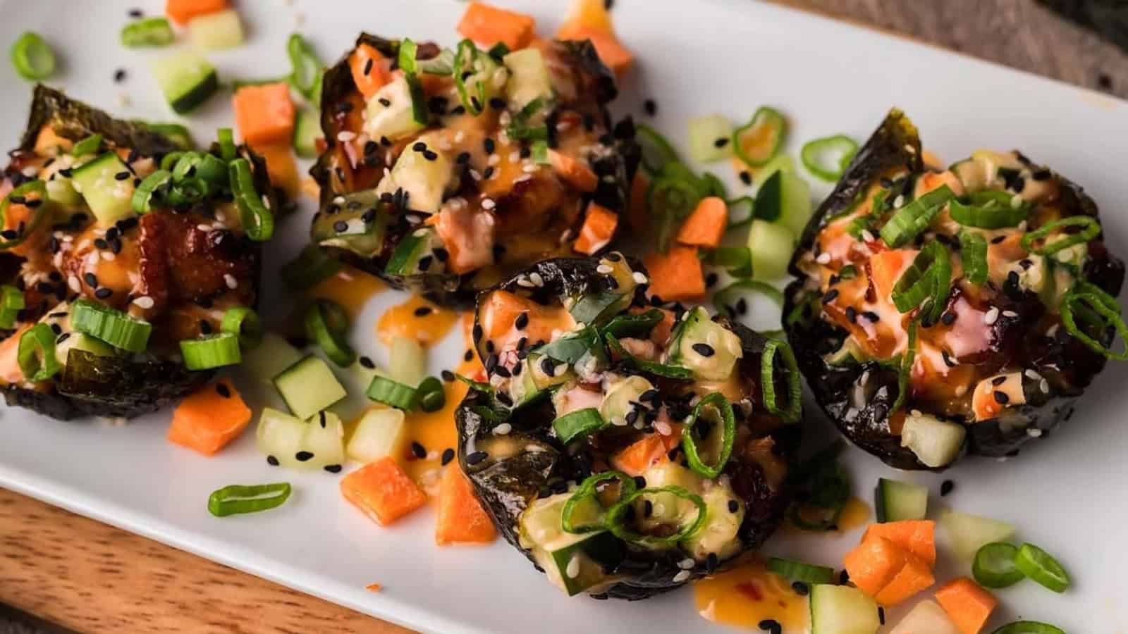 Four sushi bites topped with sauce, diced vegetables, green onions, and sesame seeds on a white plate.