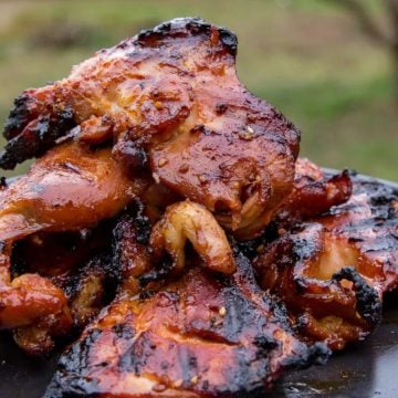 Grilled chicken pieces with a charred, glazed surface stacked on a black plate outdoors.