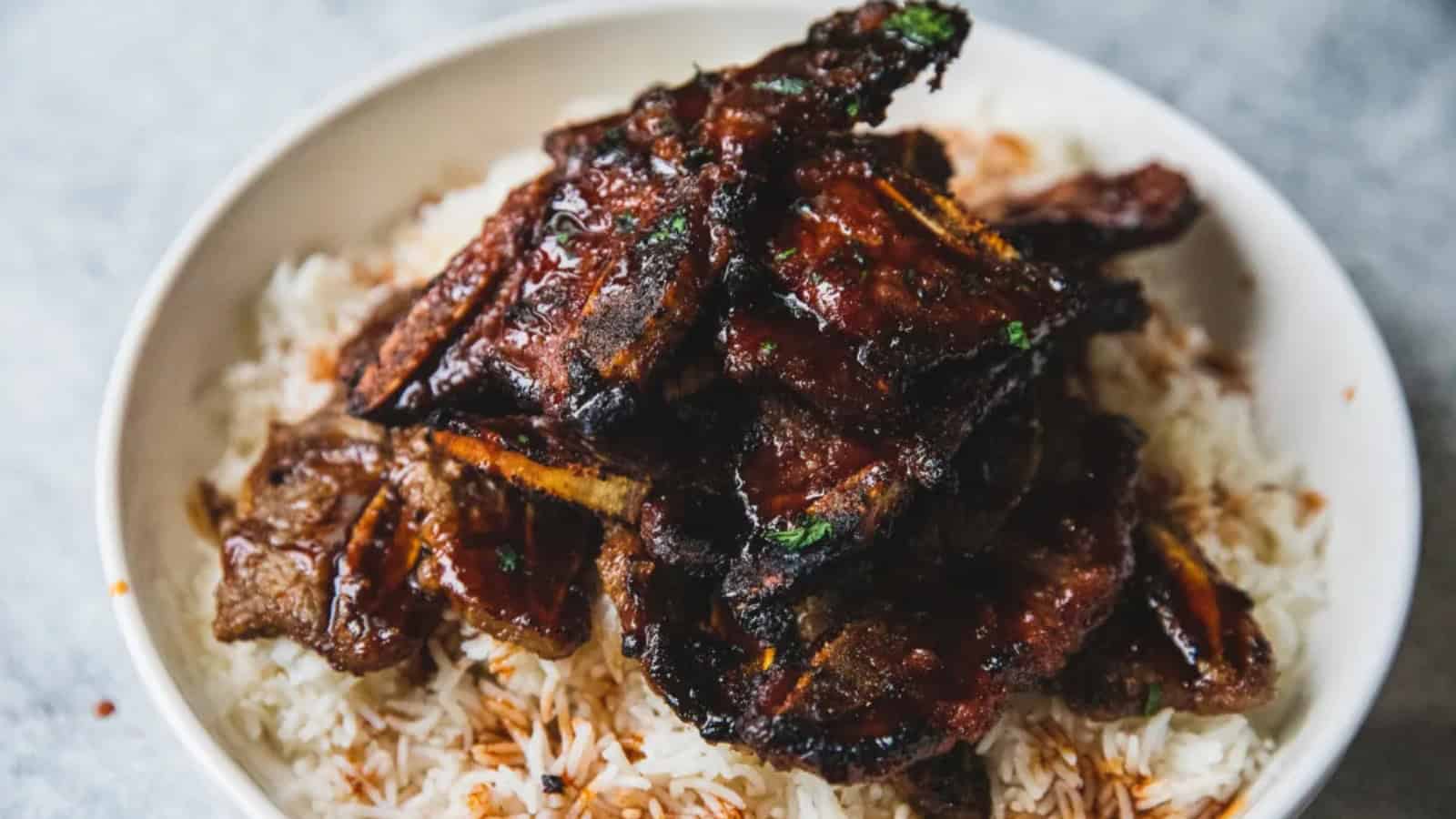 A bowl of white rice topped with glazed, grilled beef short ribs, garnished with herbs.