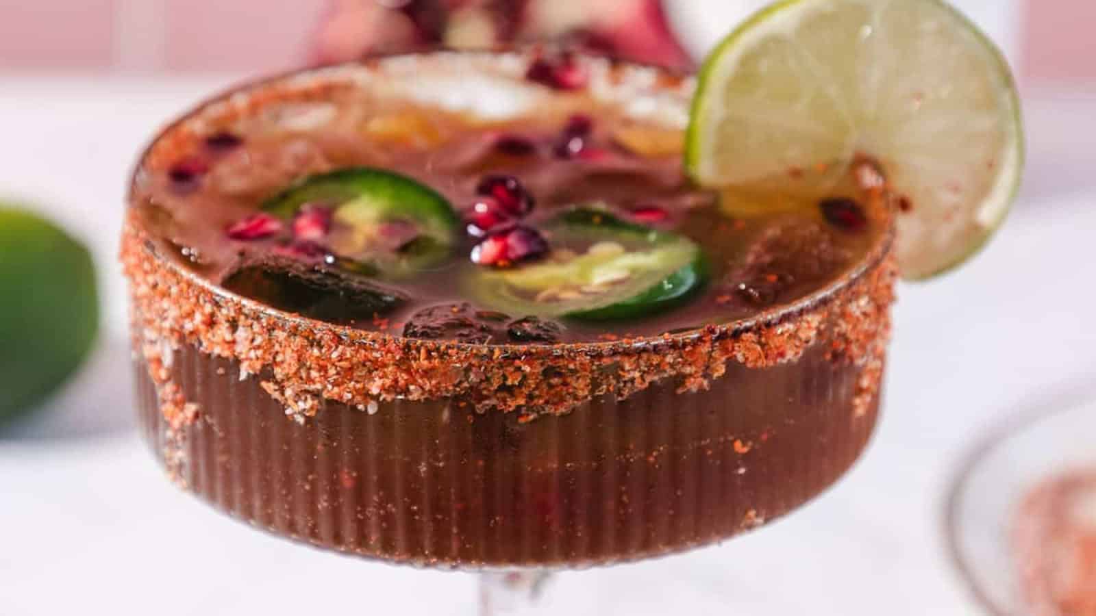 A cocktail in a salt-rimmed glass garnished with lime, jalapeño slices, and pomegranate seeds.