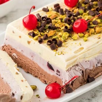 Layered ice cream cake topped with cherries, chocolate curls, and chopped pistachios on a white platter.