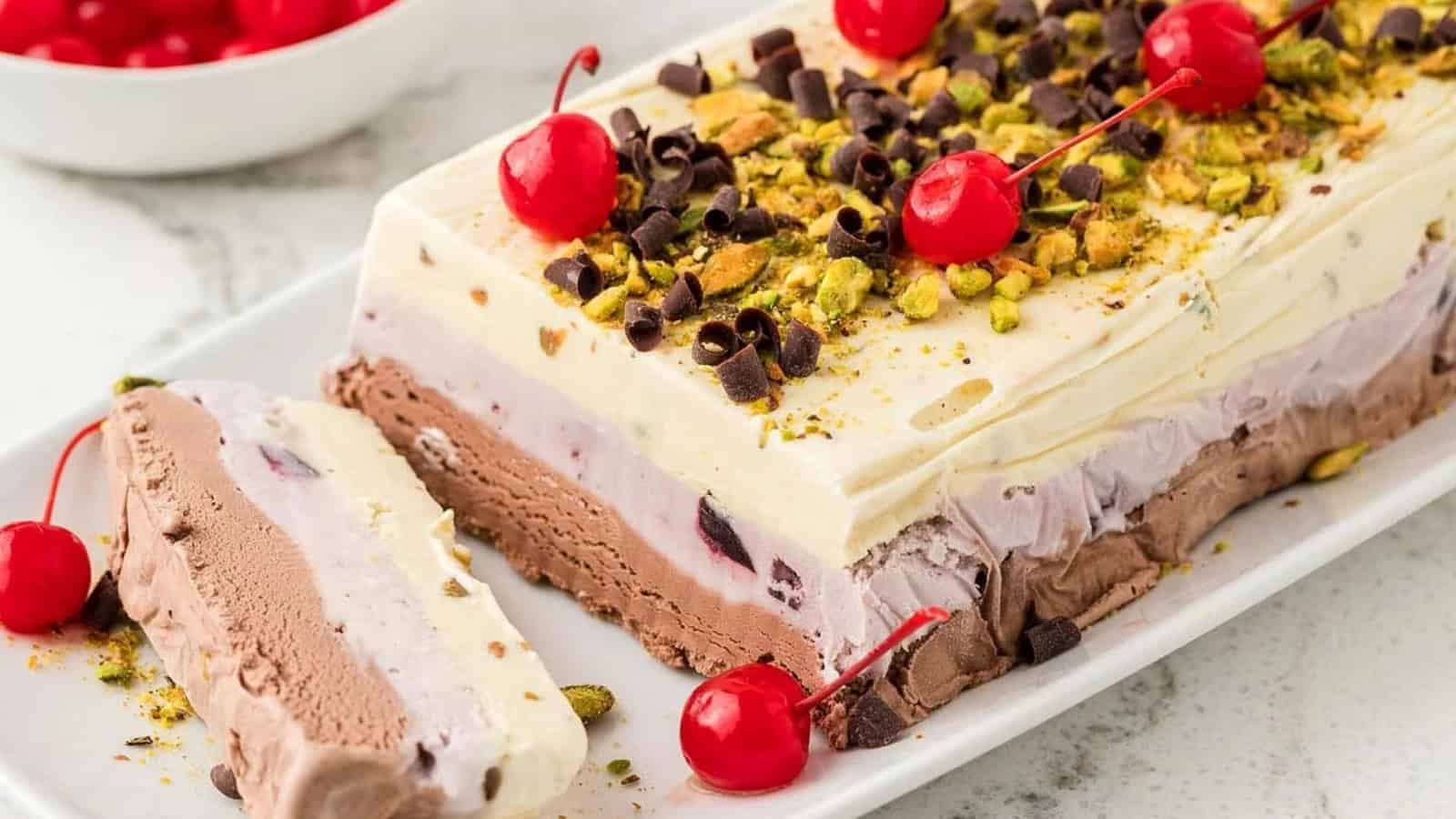 Layered ice cream cake topped with cherries, chocolate curls, and chopped pistachios on a white platter.