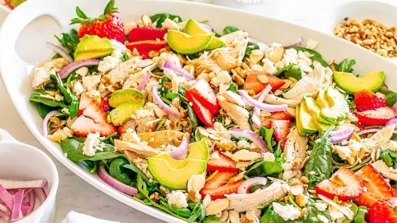 Colorful salad with spinach, strawberries, avocado, chicken, red onion, feta, and sliced almonds in a white dish.