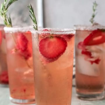 Four glasses of strawberry drinks with ice, garnished with rosemary sprigs on a light table.