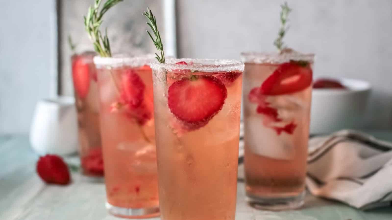 Four glasses of strawberry drinks with ice, garnished with rosemary sprigs on a light table.