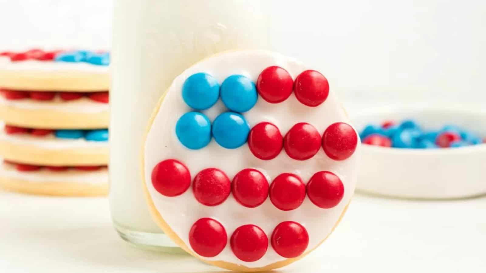 Sugar cookie decorated like an American flag with blue and red candies, in front of a glass of milk.
