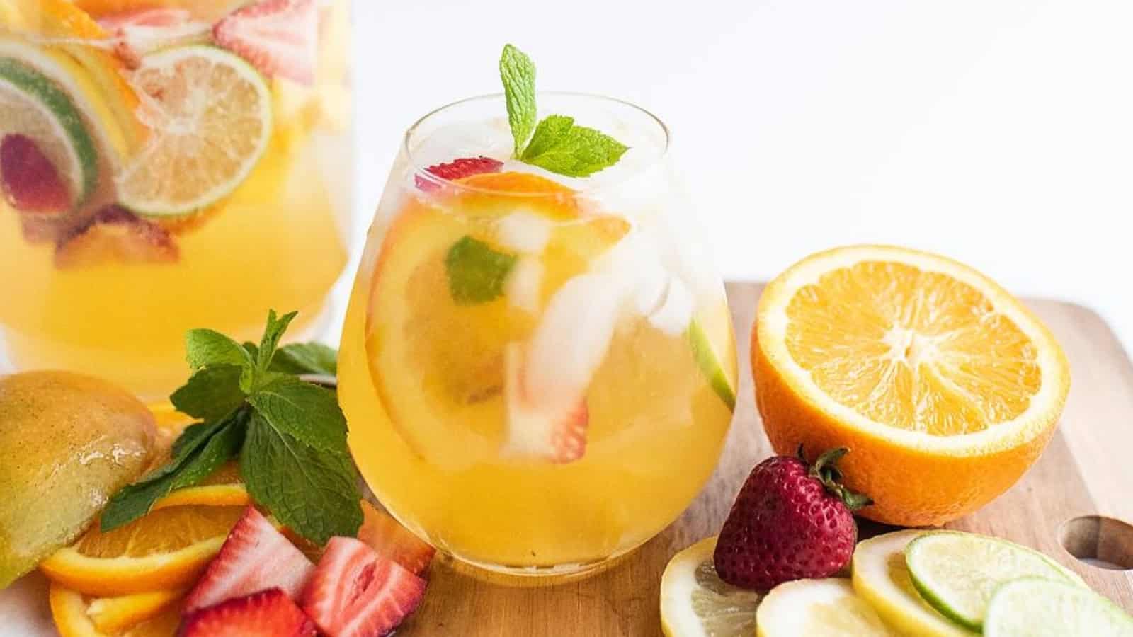 Glass of citrus fruit punch with ice, mint, and sliced fruits, surrounded by fresh fruit on a wooden board.