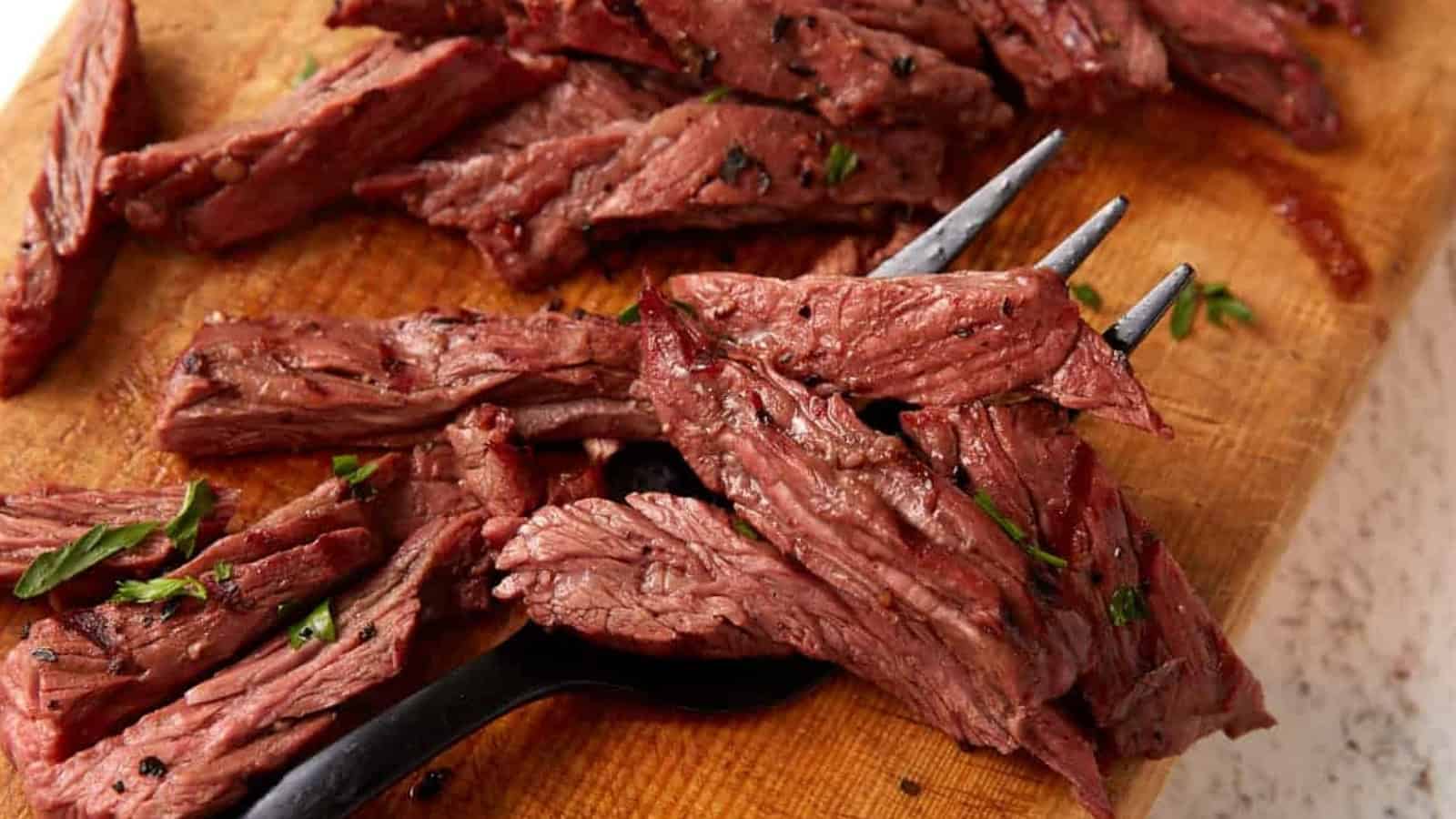 Sliced grilled steak on a wooden cutting board with a fork and some herbs scattered around.