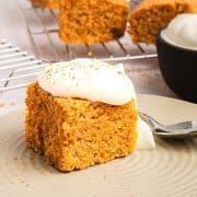 A slice of pumpkin cake with whipped cream on top sits on a plate with a spoon beside it.