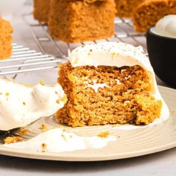 A slice of spiced cake with creamy frosting on a plate, with more cake and a bowl of frosting in the background.