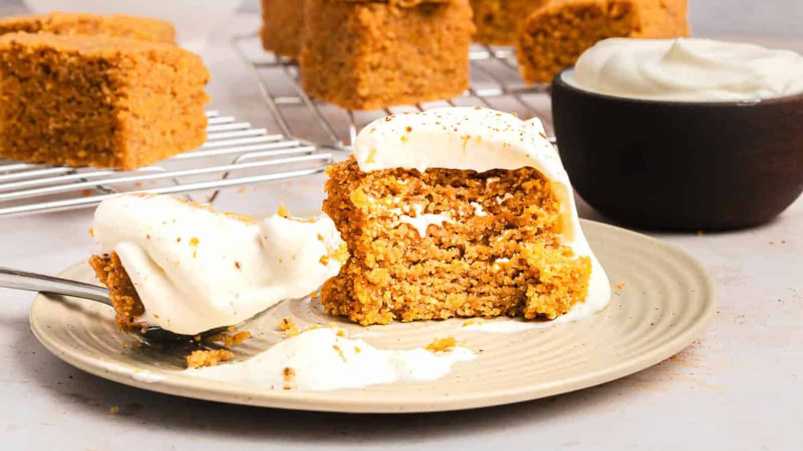 A slice of spiced cake with creamy frosting on a plate, with more cake and a bowl of frosting in the background.