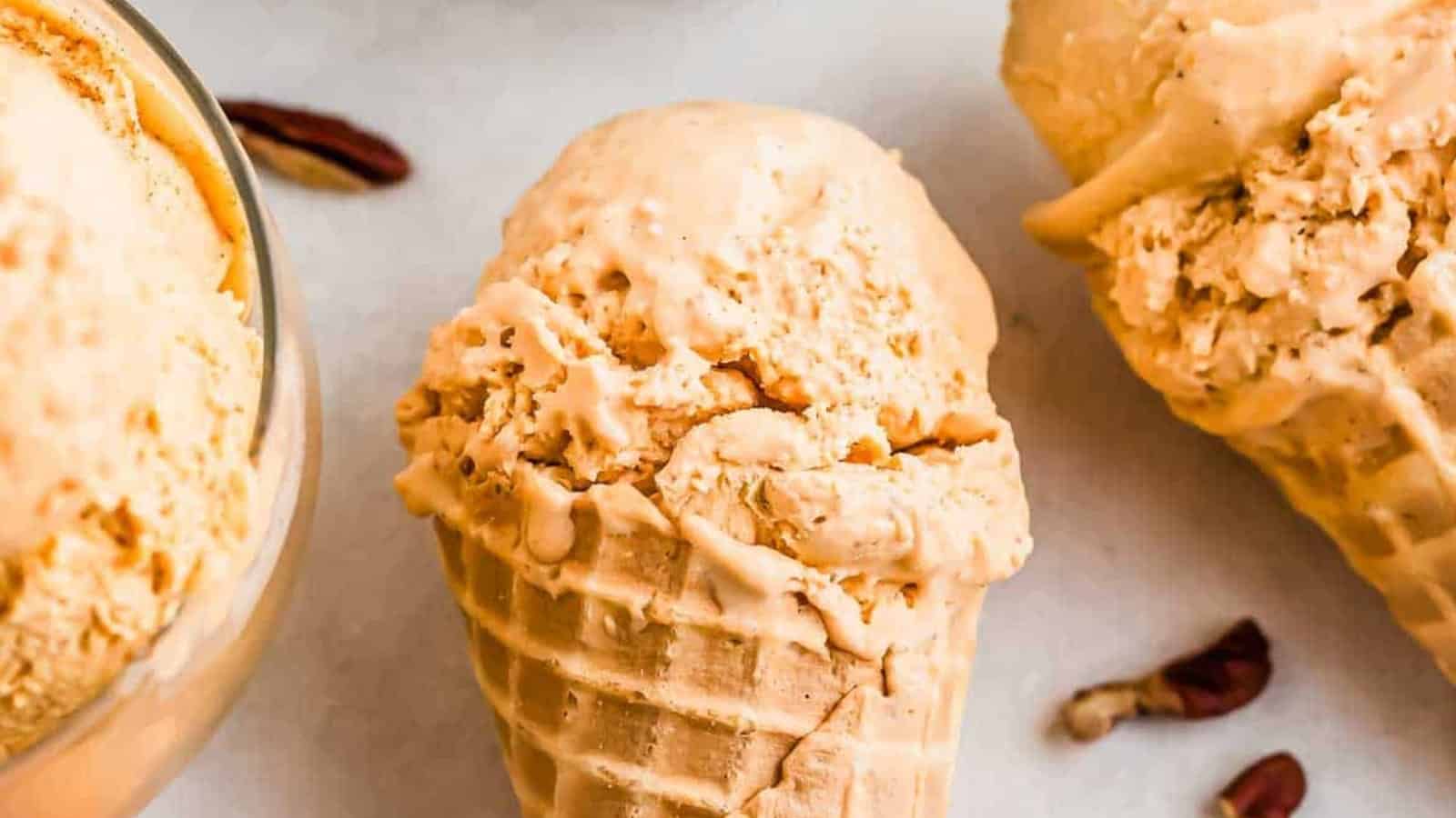Close-up of creamy, light brown ice cream scoops in waffle cones, with pecans nearby on a light surface.