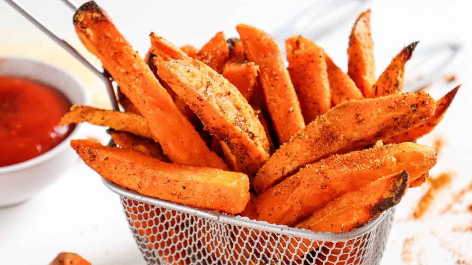 Seasoned sweet potato fries in a metal basket, with a bowl of ketchup in the background.
