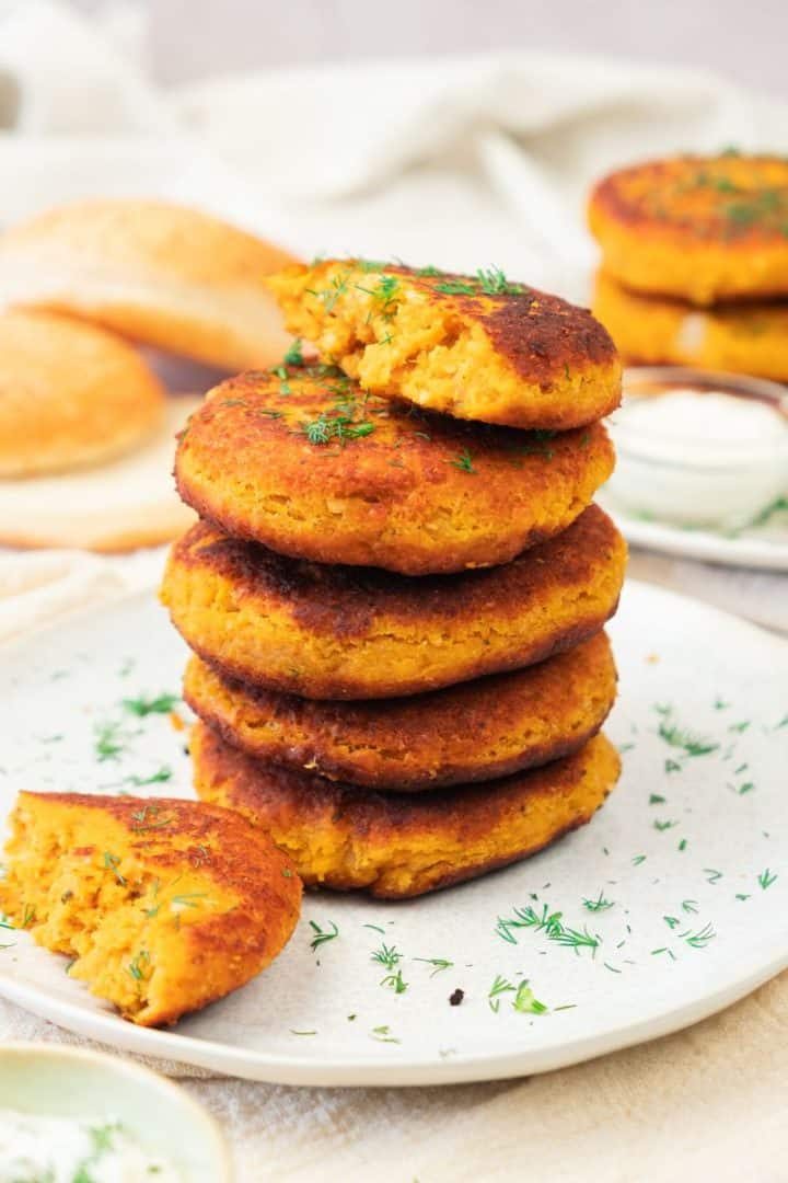 A stack of golden, crispy sweet potato and red lentil burgers with herbs on a plate; one patty is broken to show the delicious inside.