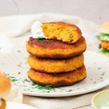 Three golden chickpea patties, inspired by sweet potato and red lentil burgers, are stacked on a plate, topped with a half patty and a dollop of white sauce.