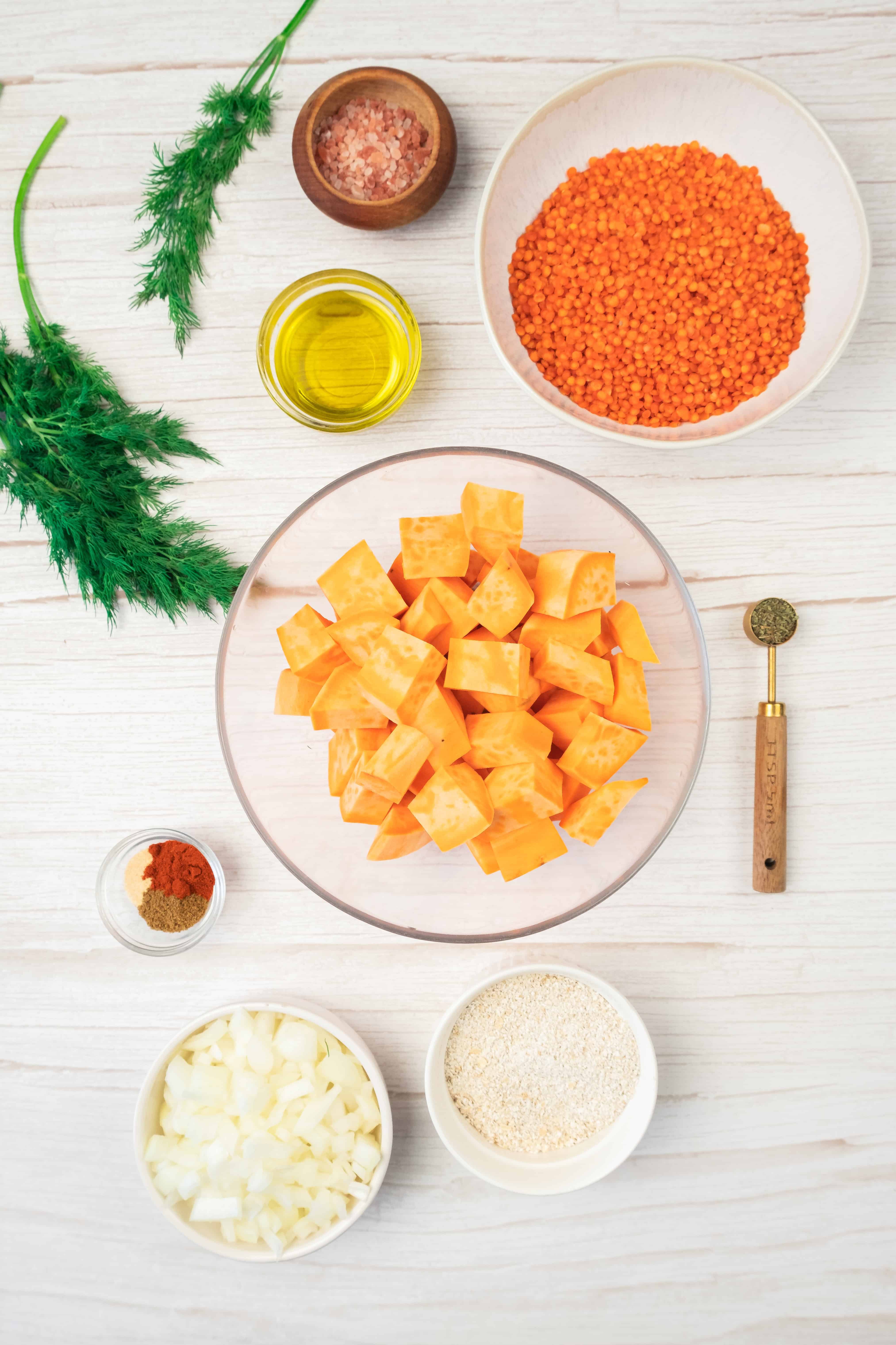 Top view of ingredients for sweet potato and red lentil burgers, including chopped sweet potatoes, red lentils, oil, onions, spices, and herbs arranged on a white surface.