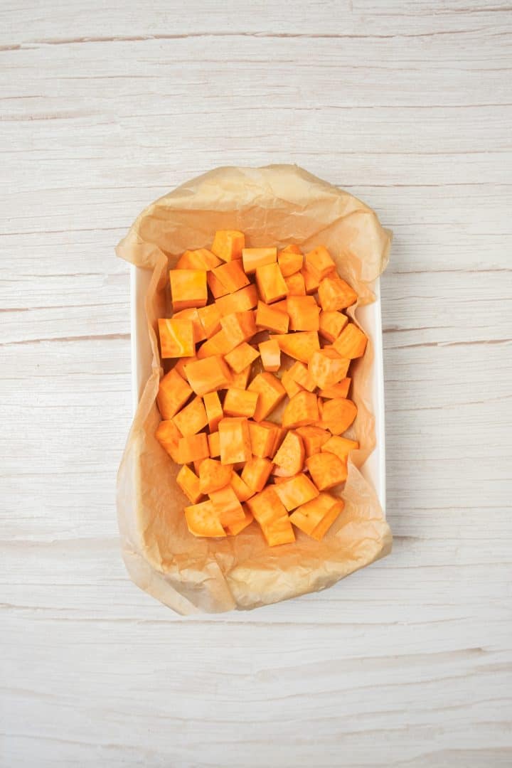 Cubed sweet potatoes on parchment paper in a rectangular white dish, perfect for making sweet potato and red lentil burgers, sit atop a light wooden surface.