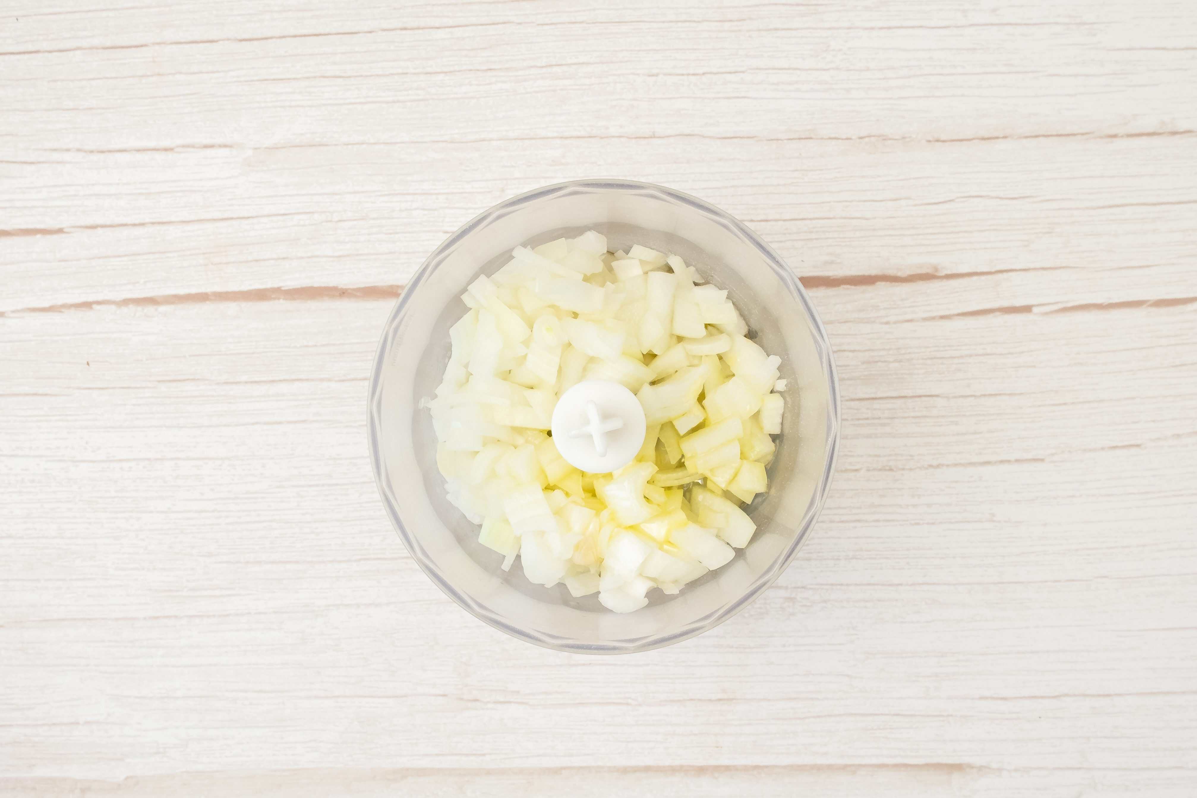 Chopped onions in a food processor bowl on a light wooden surface, viewed from above, ready to be mixed into sweet potato and red lentil burgers.