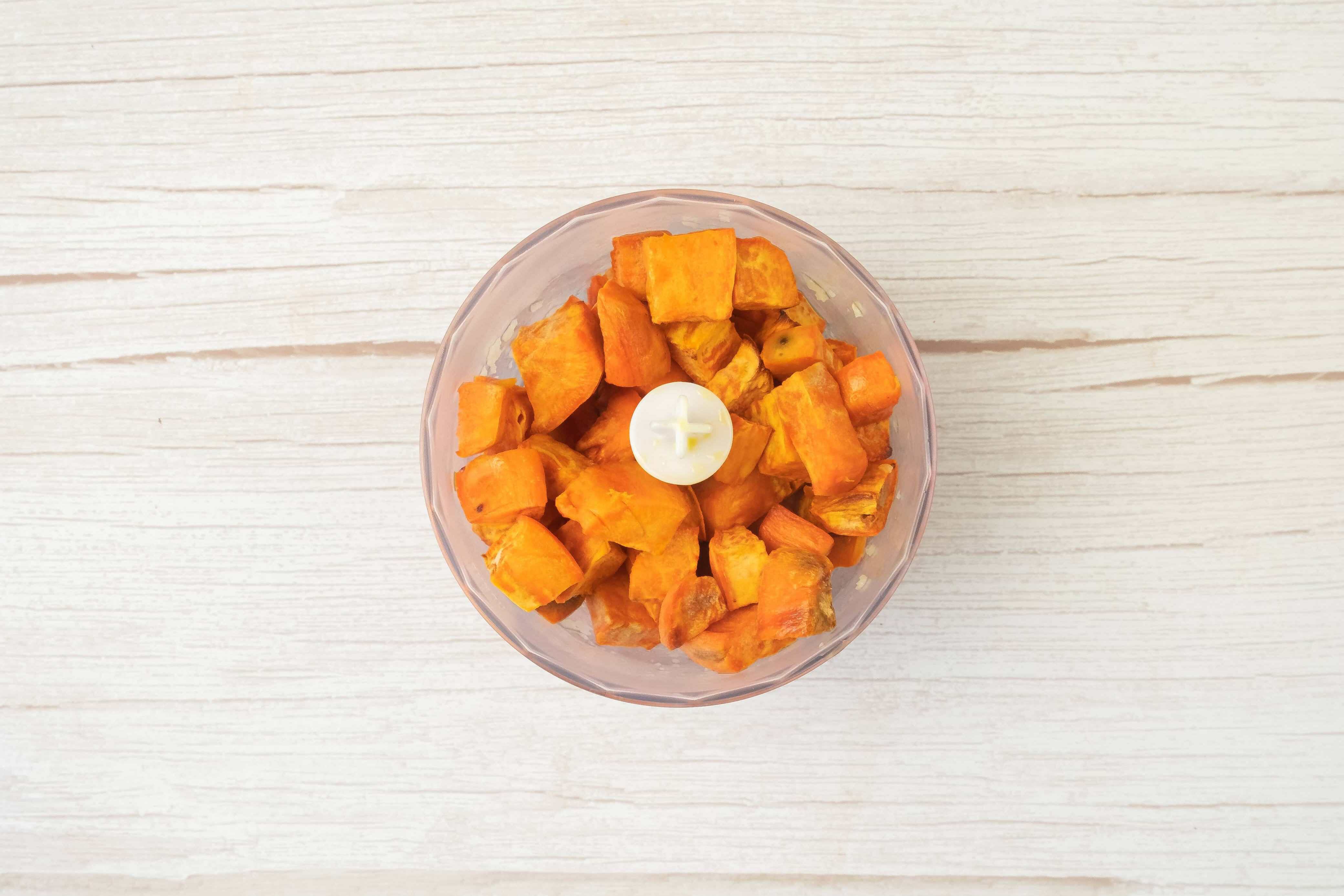Cubed orange sweet potato pieces in a food processor bowl, ready to be blended for homemade sweet potato and red lentil burgers, on a light wooden surface.