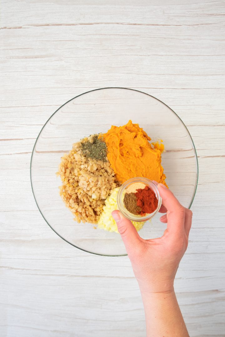 A hand adds spices to a bowl with mashed sweet potato, chopped onions, rice, and herbs-ingredients coming together for tasty sweet potato and red lentil burgers on a light surface.