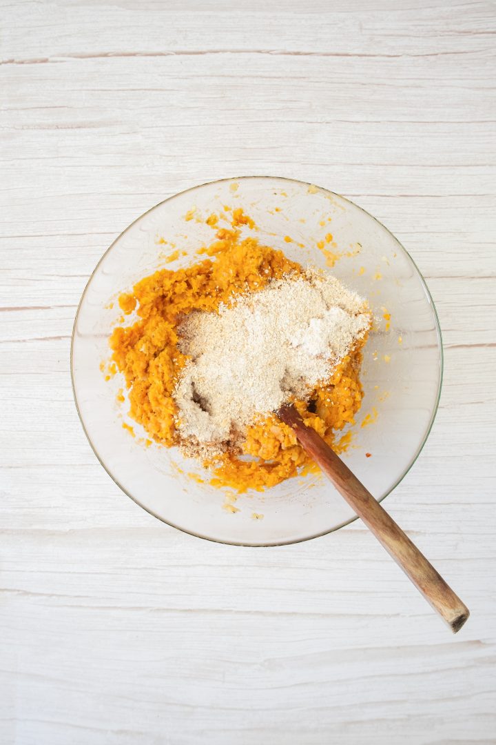 A glass bowl with mashed orange mixture and breadcrumbs, perfect for making sweet potato and red lentil burgers, stirred with a wooden spoon on a light surface.