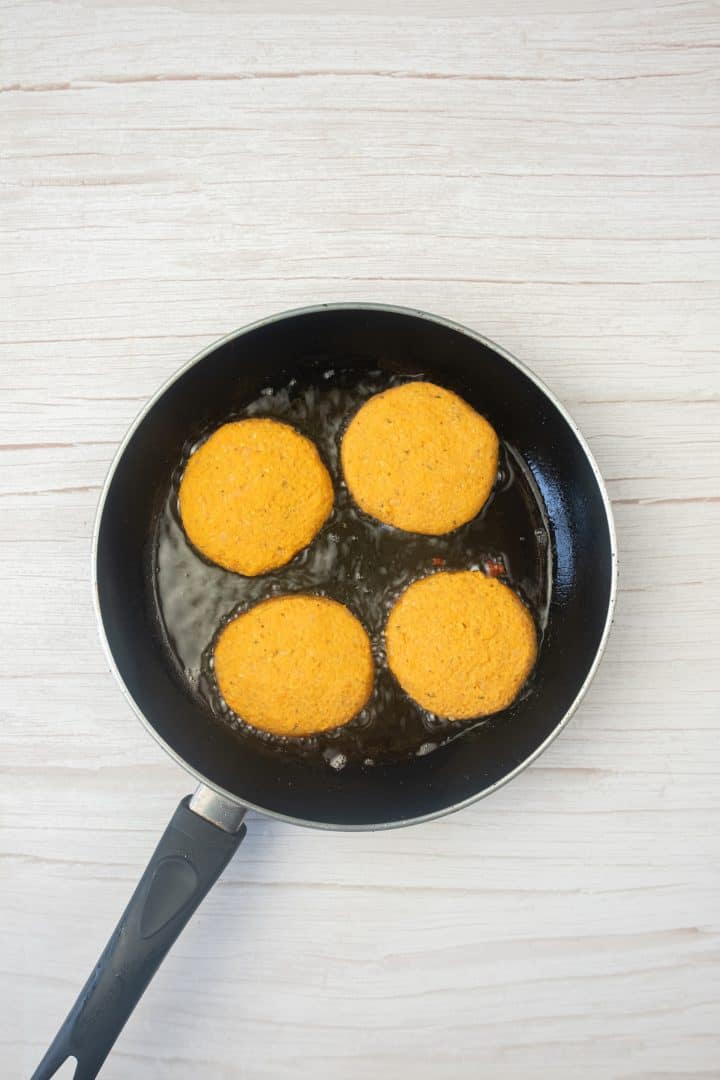 Four sweet potato and red lentil burgers frying in oil in a black pan on a light-colored wooden surface.