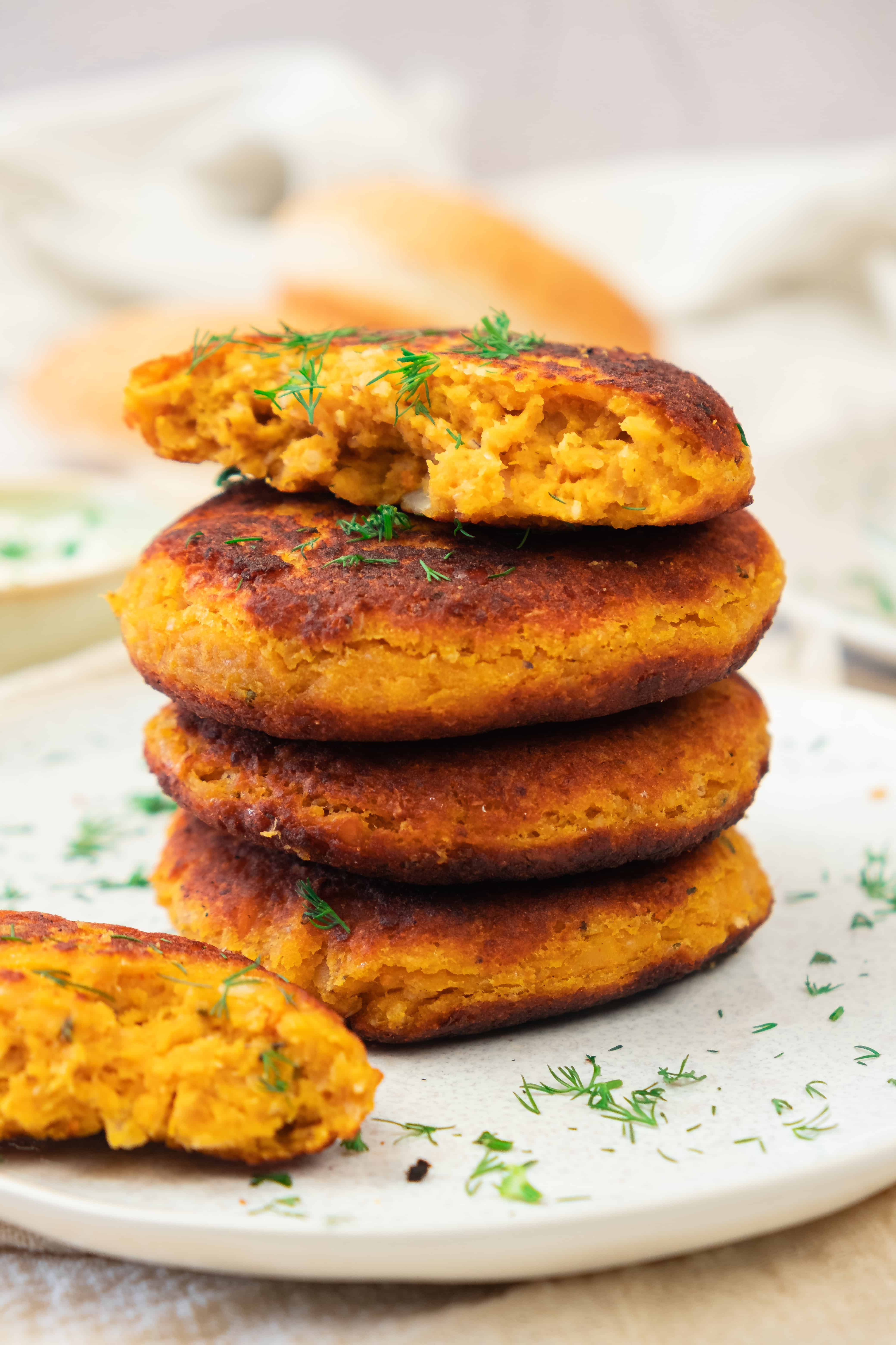 Four golden-brown sweet potato and red lentil burgers stacked on a plate, garnished with fresh dill, one patty partially eaten.