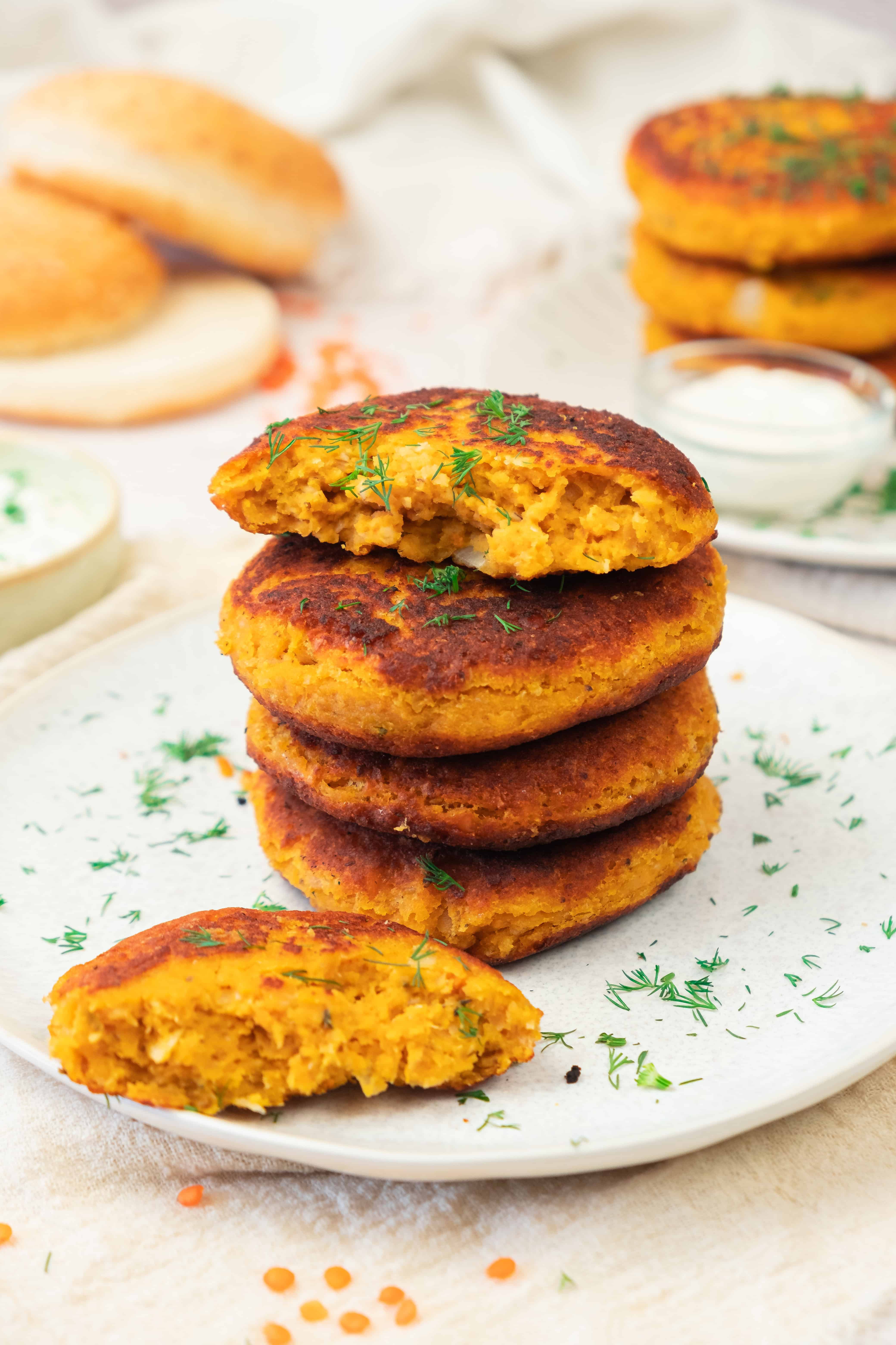 A stack of golden-brown sweet potato and red lentil burgers on a plate, garnished with chopped herbs, with one patty broken open.