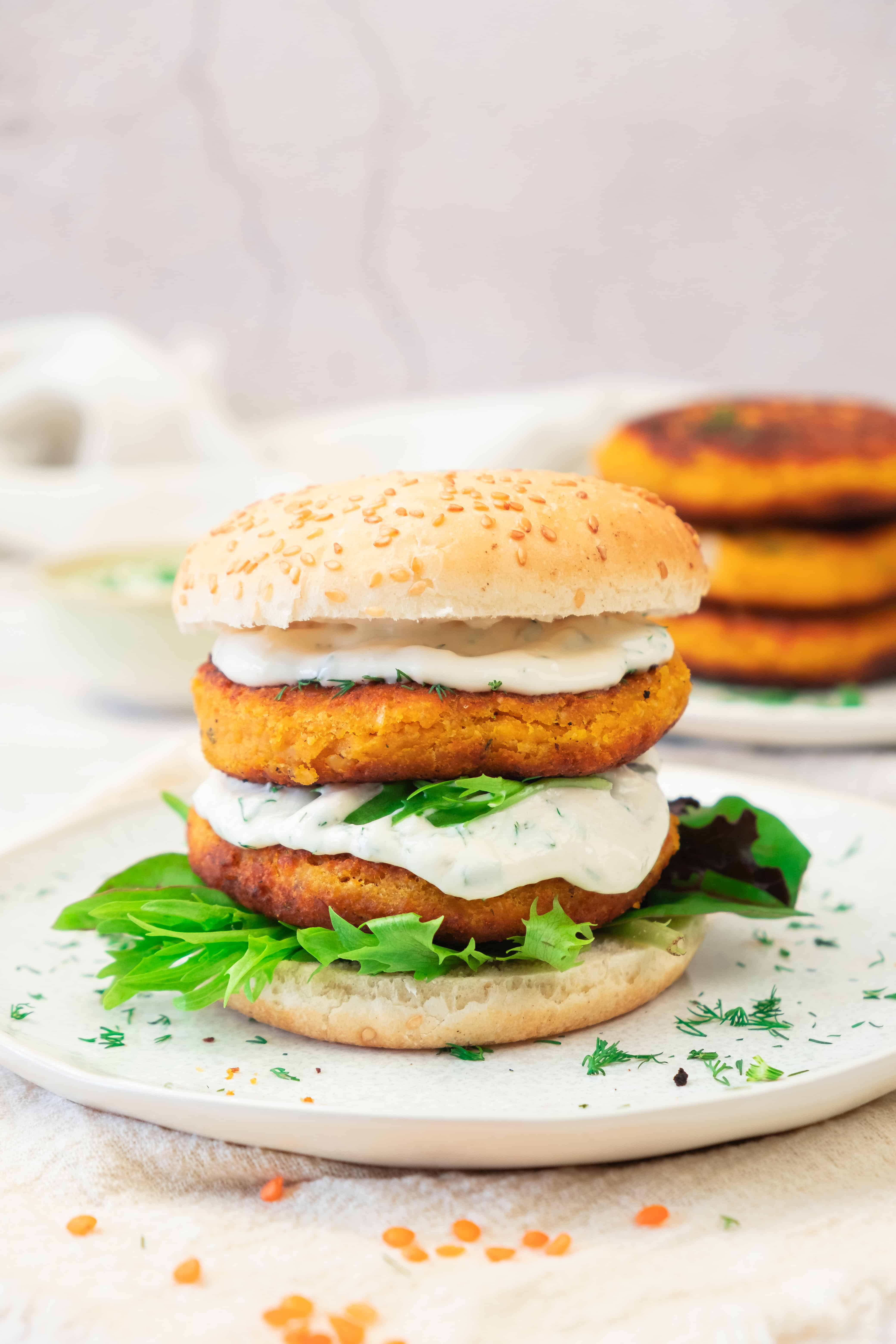 A sweet potato and red lentil burger with leafy greens and creamy sauce in a sesame bun, served on a white plate.