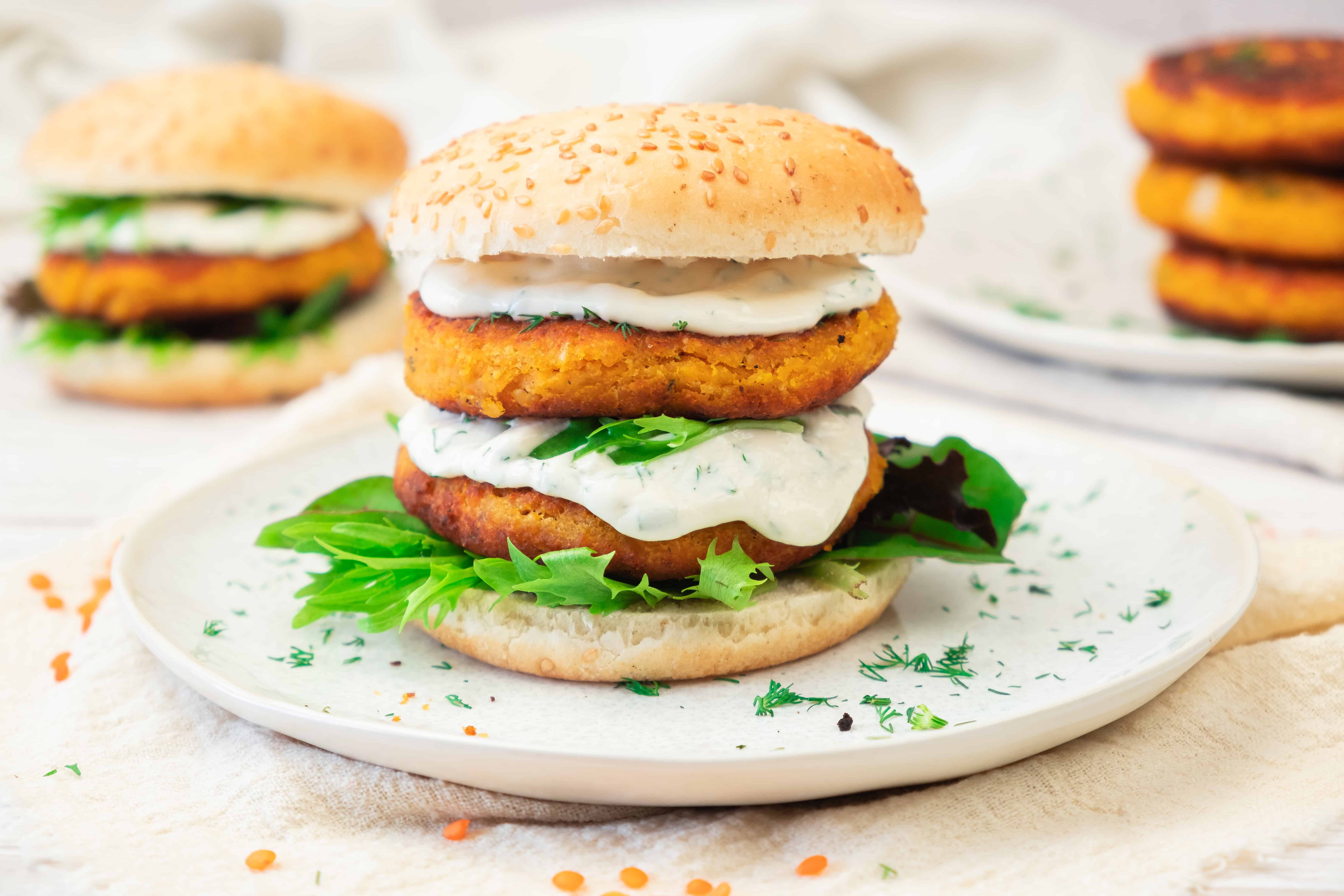 A sweet potato and red lentil veggie burger with lettuce and creamy sauce on a sesame bun, served on a white plate.