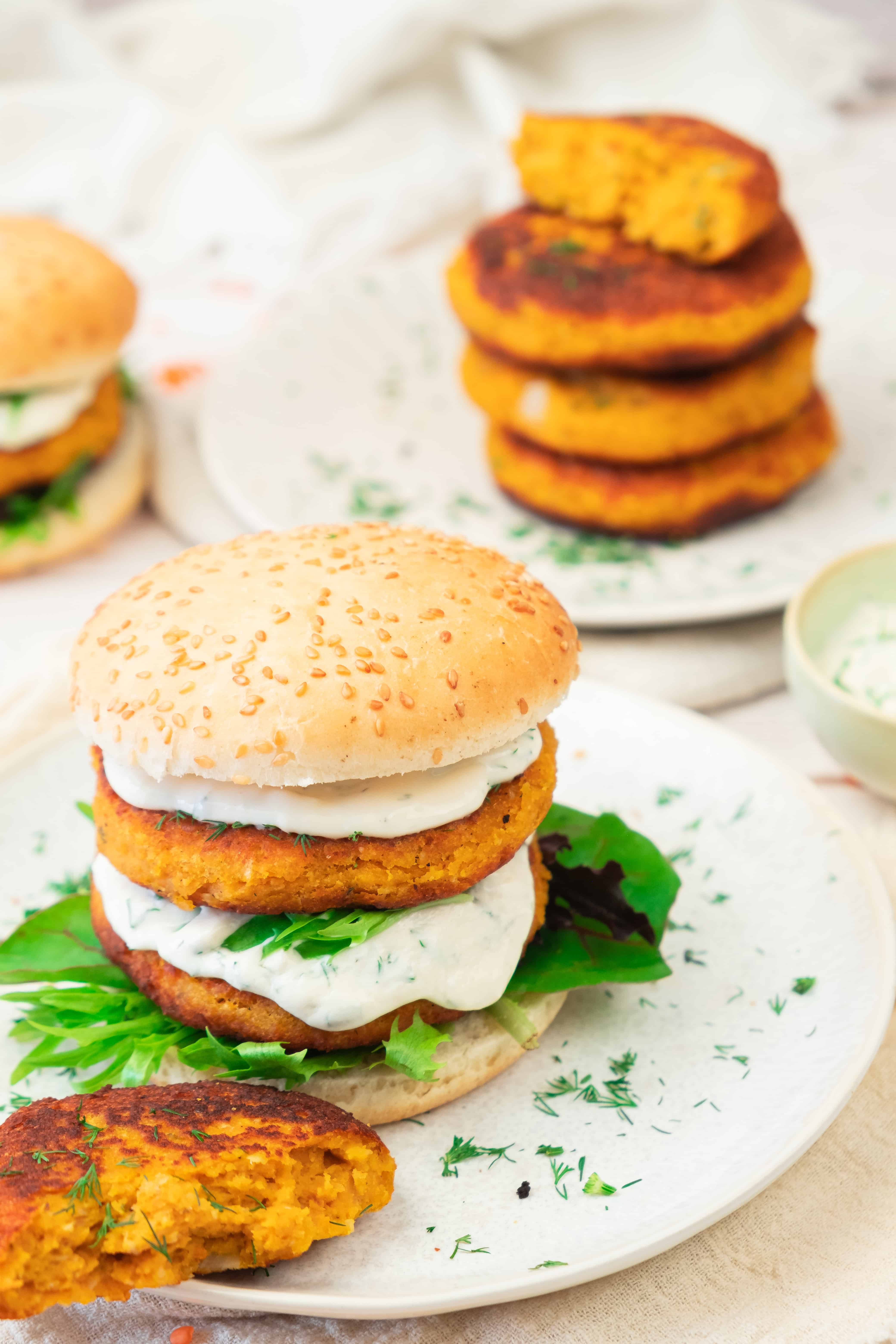 A veggie burger with leafy greens and creamy sauce on a sesame bun, featuring sweet potato and red lentil burgers, with extra patties stacked in the background.
