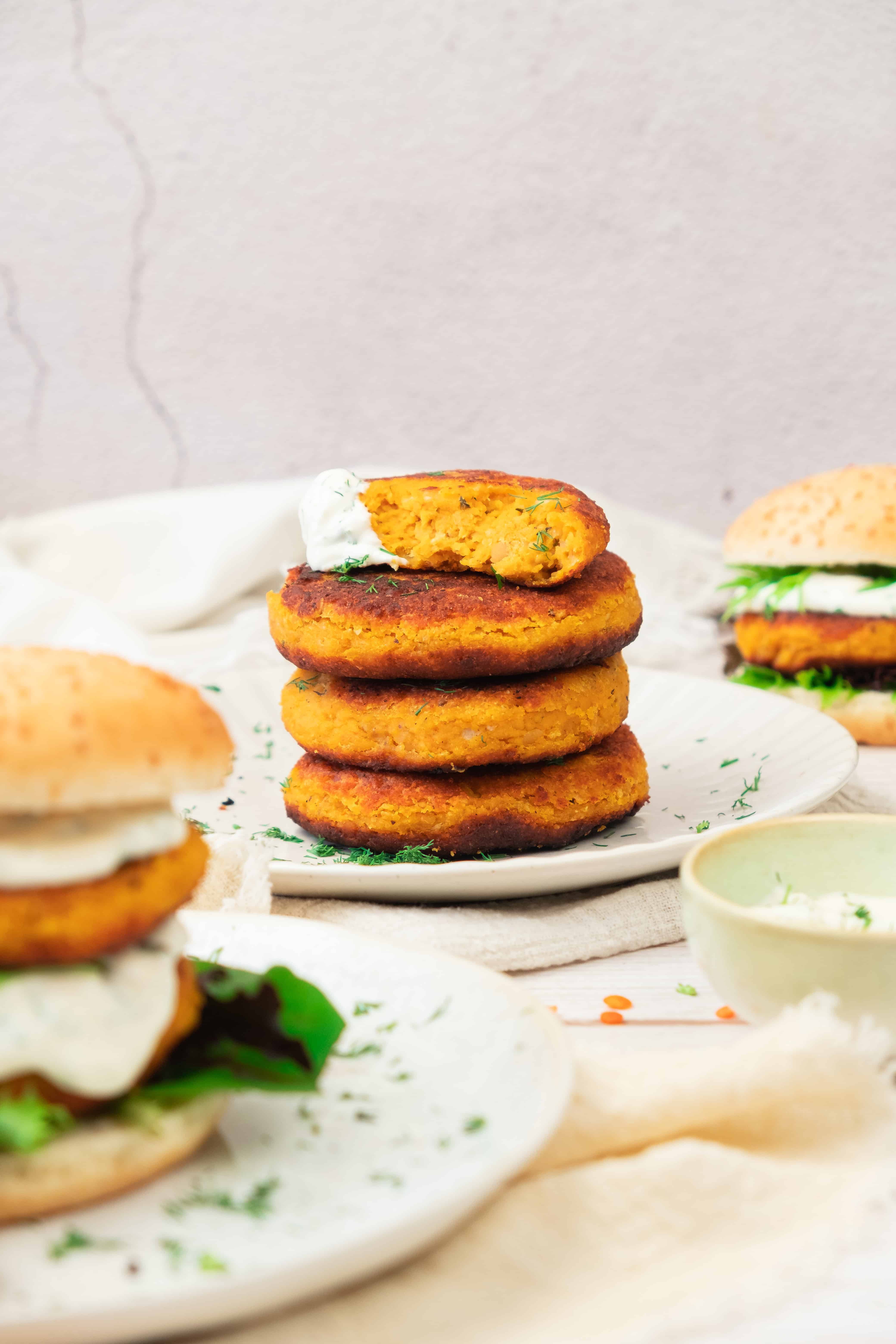 A stack of sweet potato and red lentil burgers topped with a dollop of sauce, with burgers and greens in the background.