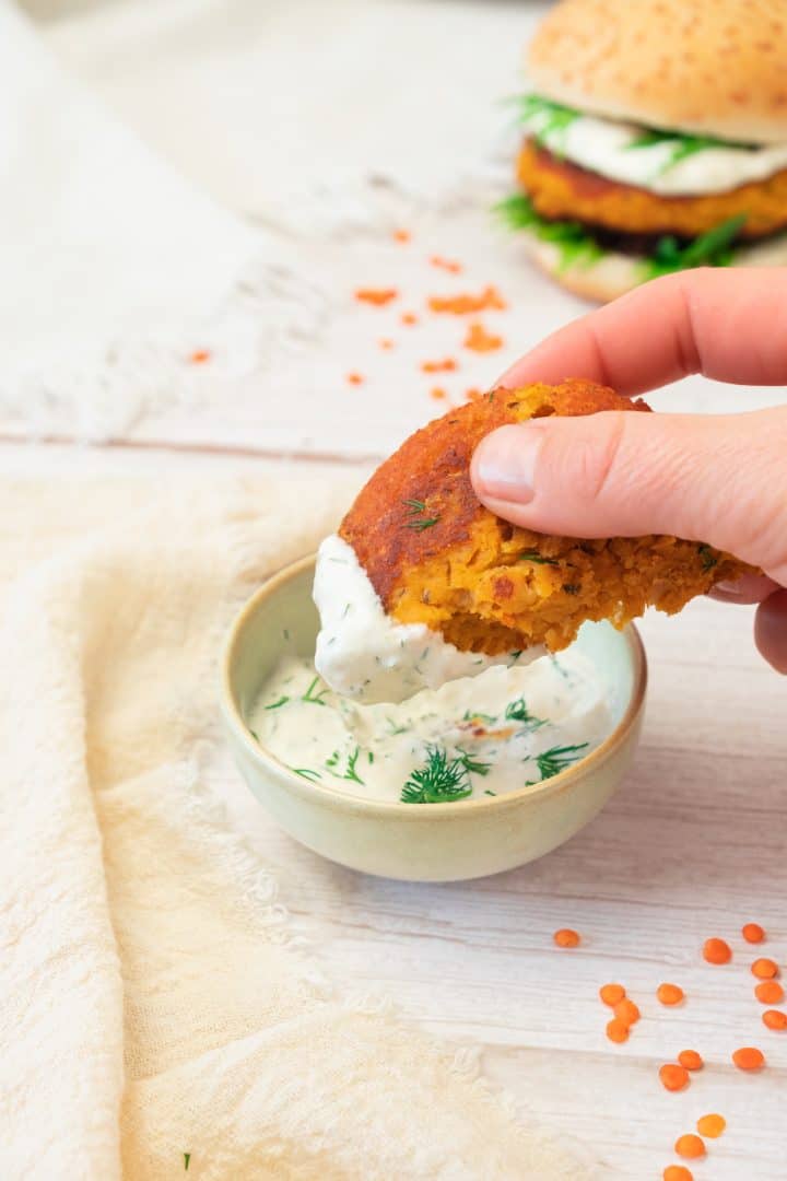 Hand dipping a veggie patty into herbed yogurt sauce, with sweet potato and red lentil burgers and whole red lentils in the background.