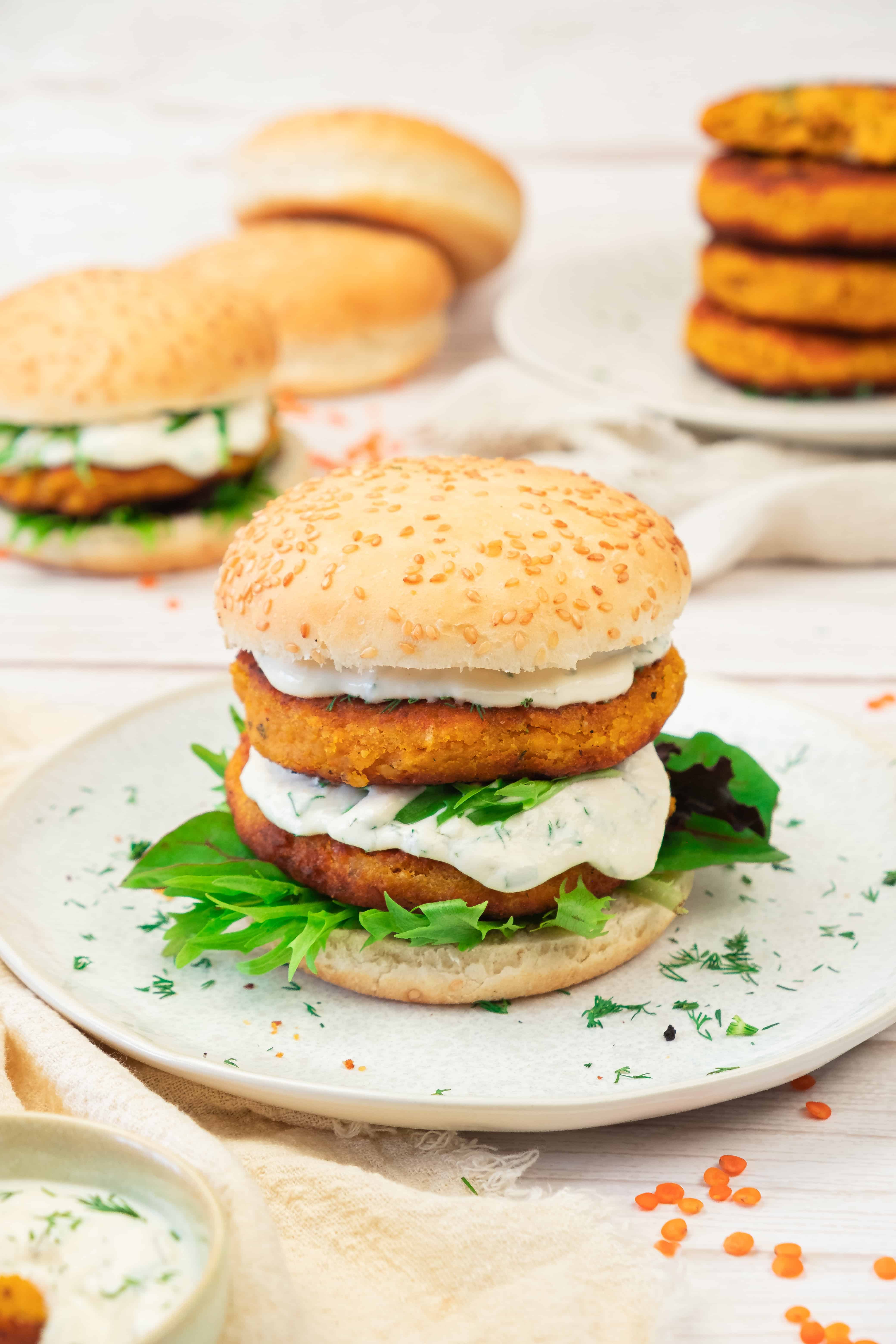 A sweet potato and red lentil burger with lettuce and creamy sauce on a sesame bun, served on a plate alongside other veggie burgers.