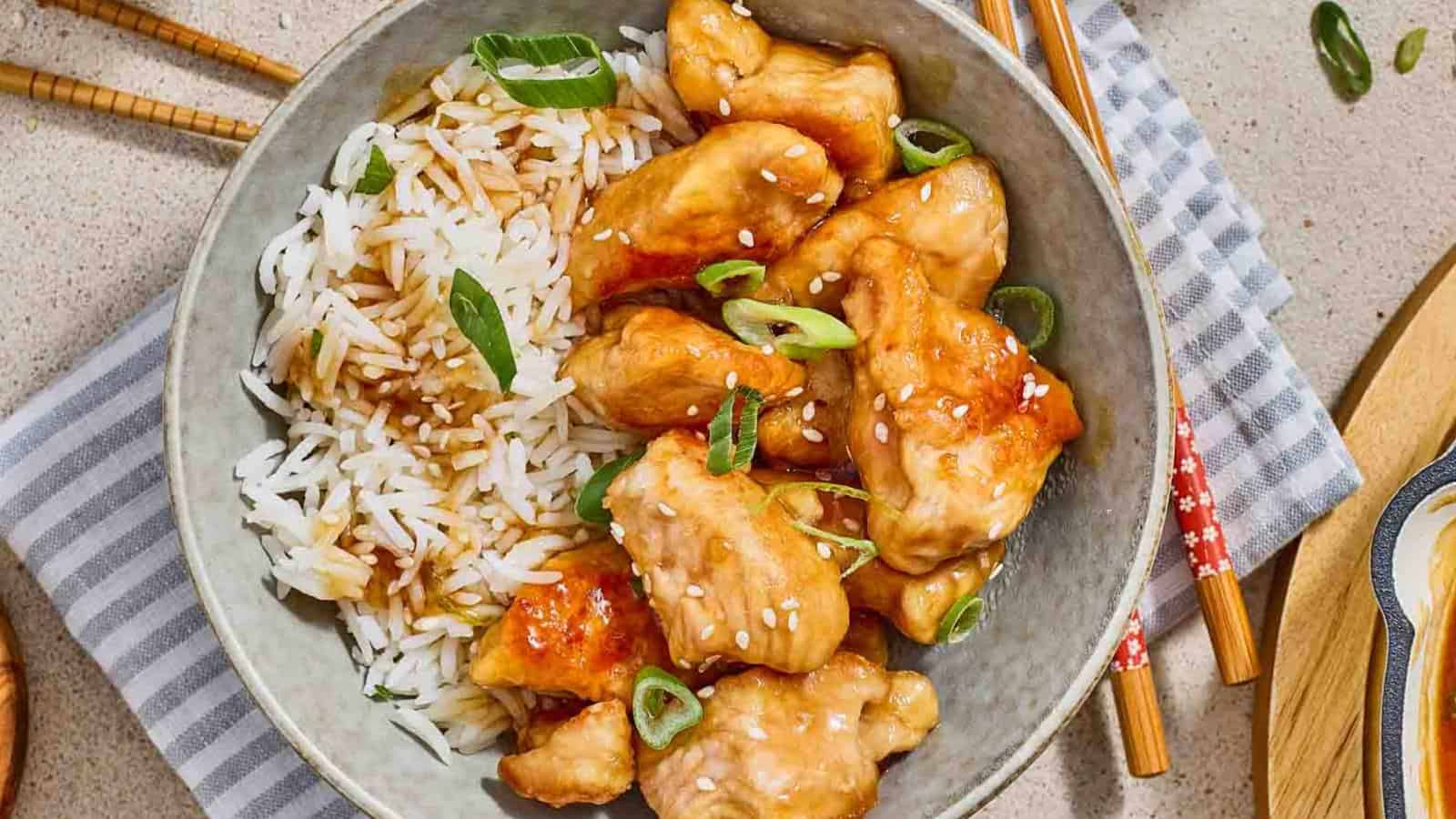 A bowl of white rice and glazed chicken pieces topped with sesame seeds and sliced green onions, with chopsticks and a striped napkin nearby&mdash;perfect for those seeking delicious budget chicken recipes.