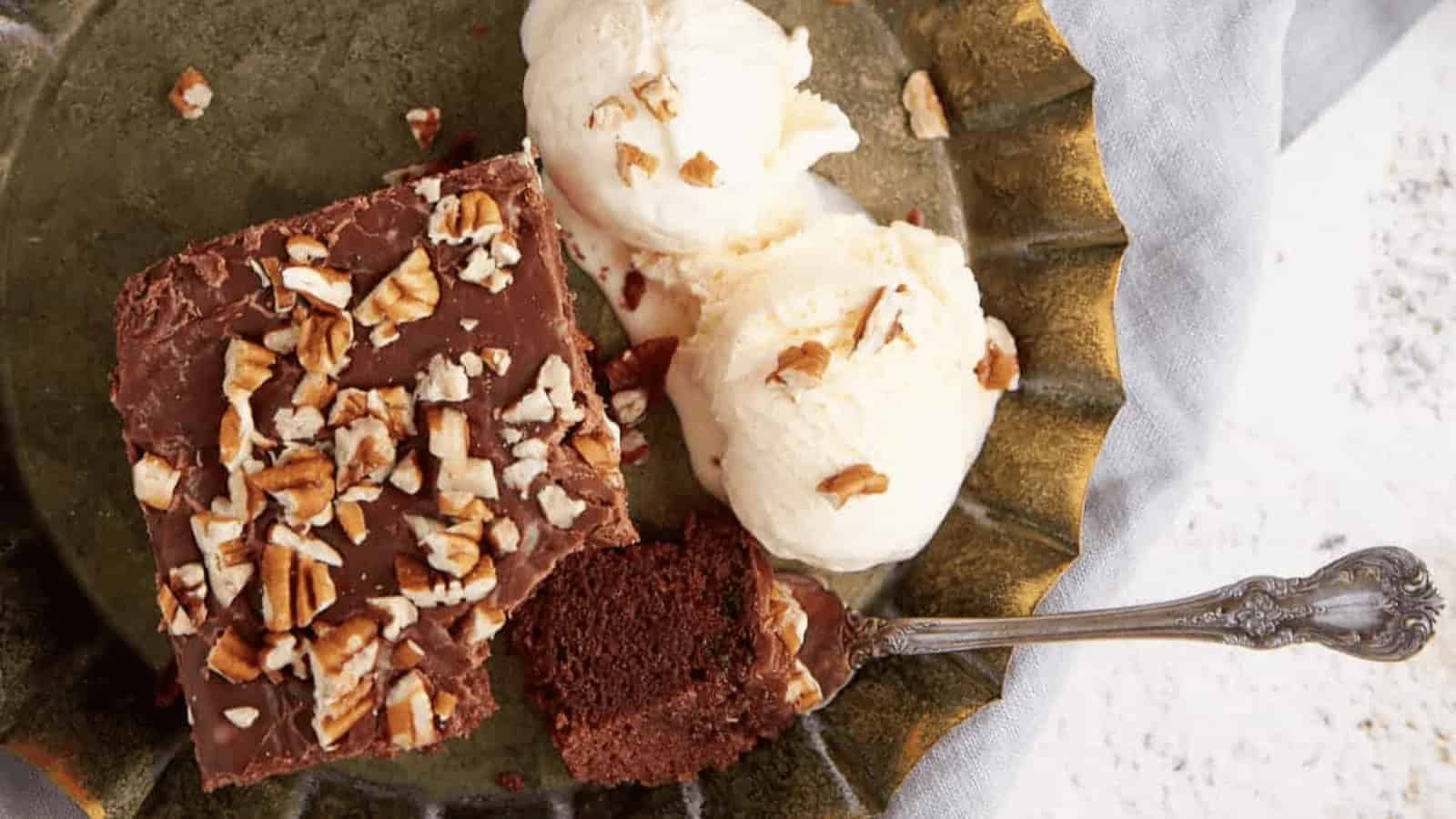 A slice of chocolate cake with nuts and two scoops of vanilla ice cream on a plate with a spoon.