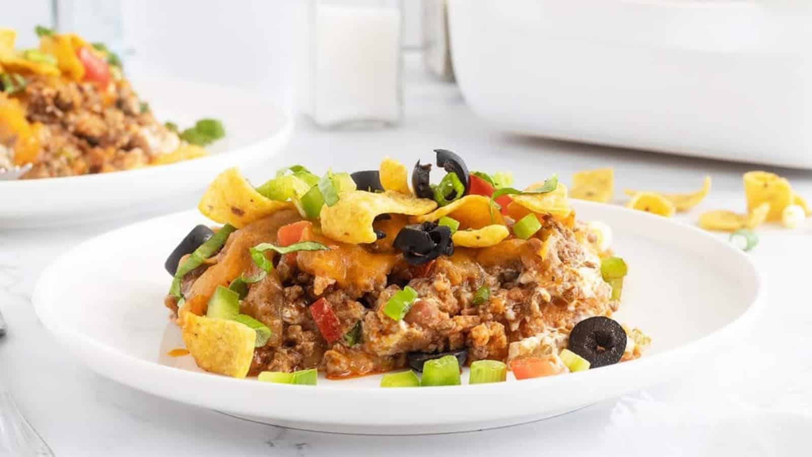 A serving of Frito pie topped with cheese, black olives, green onions, and corn chips on a white plate.
