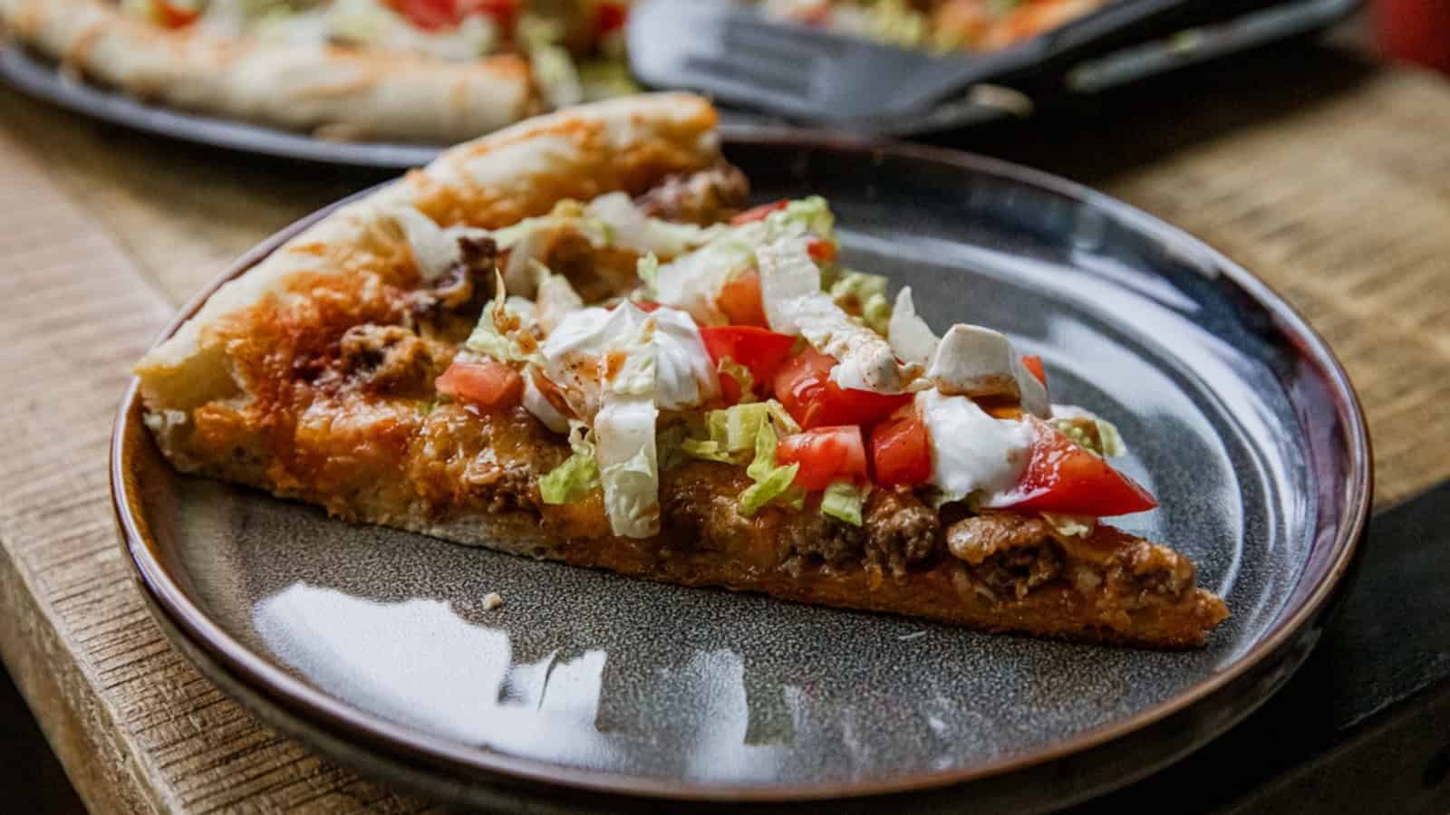 A slice of taco pizza topped with lettuce, tomatoes, and sour cream on a dark plate.