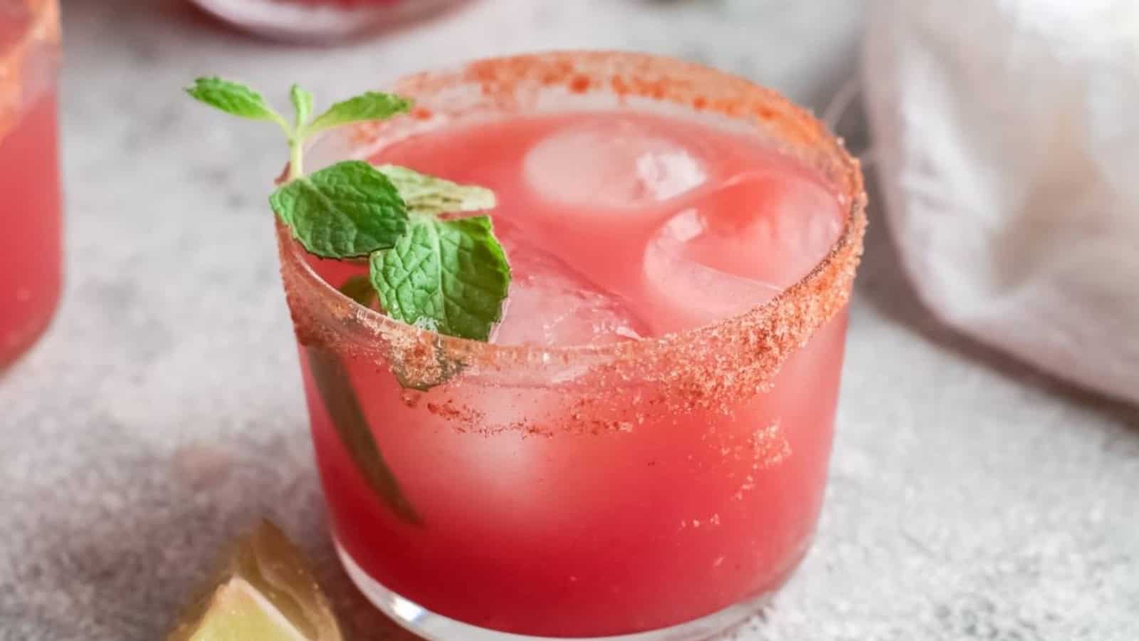 A pink cocktail with ice, mint garnish, and chili powder rim in a glass, next to a lime wedge.