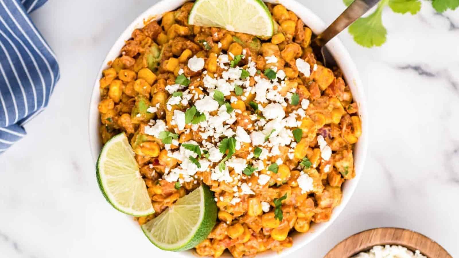 A bowl of Mexican street corn salad topped with cheese, cilantro, and lime wedges on a marble surface.