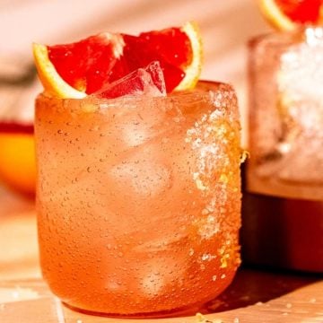 A pink cocktail with ice and grapefruit wedge, served in a salt-rimmed glass on a sunny table.