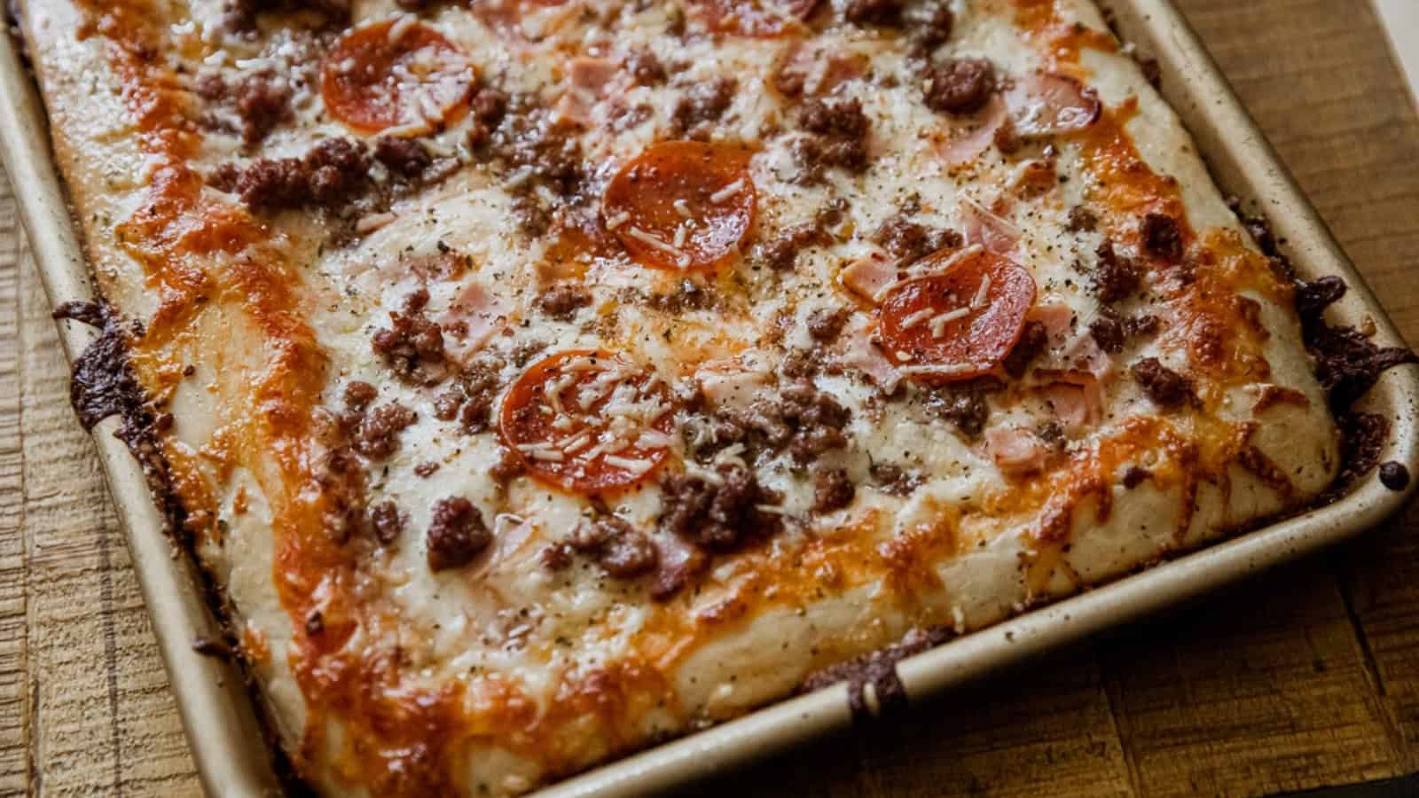 Rectangular homemade pizza in a baking pan topped with pepperoni, sausage, and melted cheese.