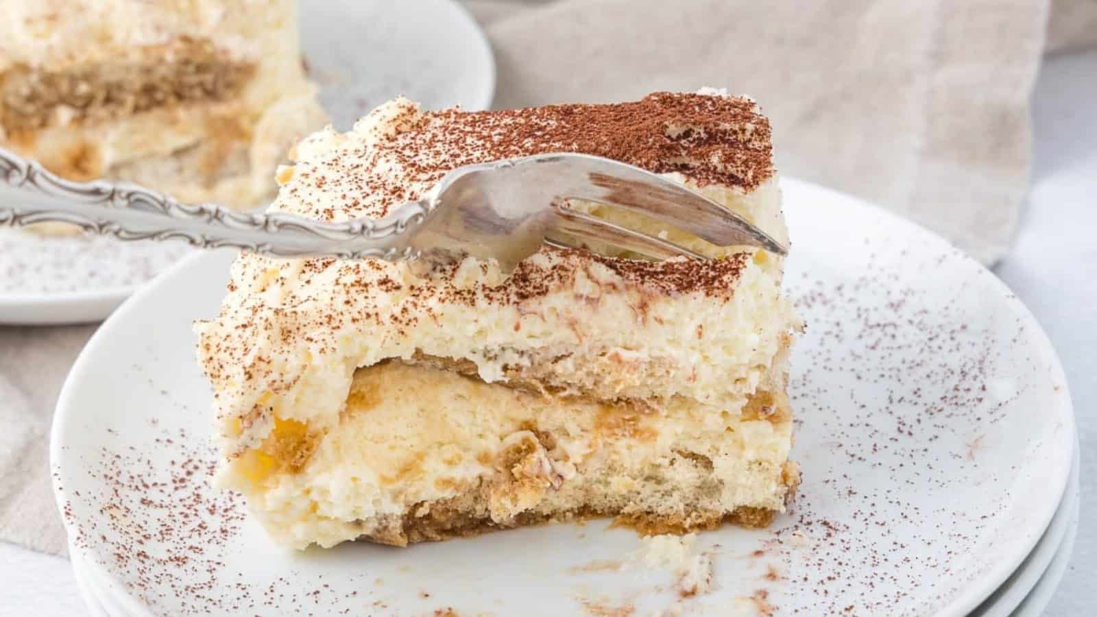 A fork slicing into a piece of tiramisu on a white plate, dusted with cocoa powder.