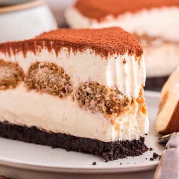 A slice of tiramisu with creamy layers and cocoa topping on a plate, with a fork and a cup in the background.