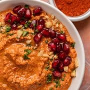 A bowl of red dip garnished with pomegranate seeds, herbs, and chopped nuts, with paprika in a small dish nearby.