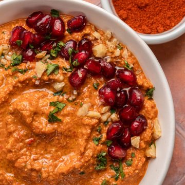 A bowl of red dip garnished with pomegranate seeds, herbs, and chopped nuts, with paprika in a small dish nearby.